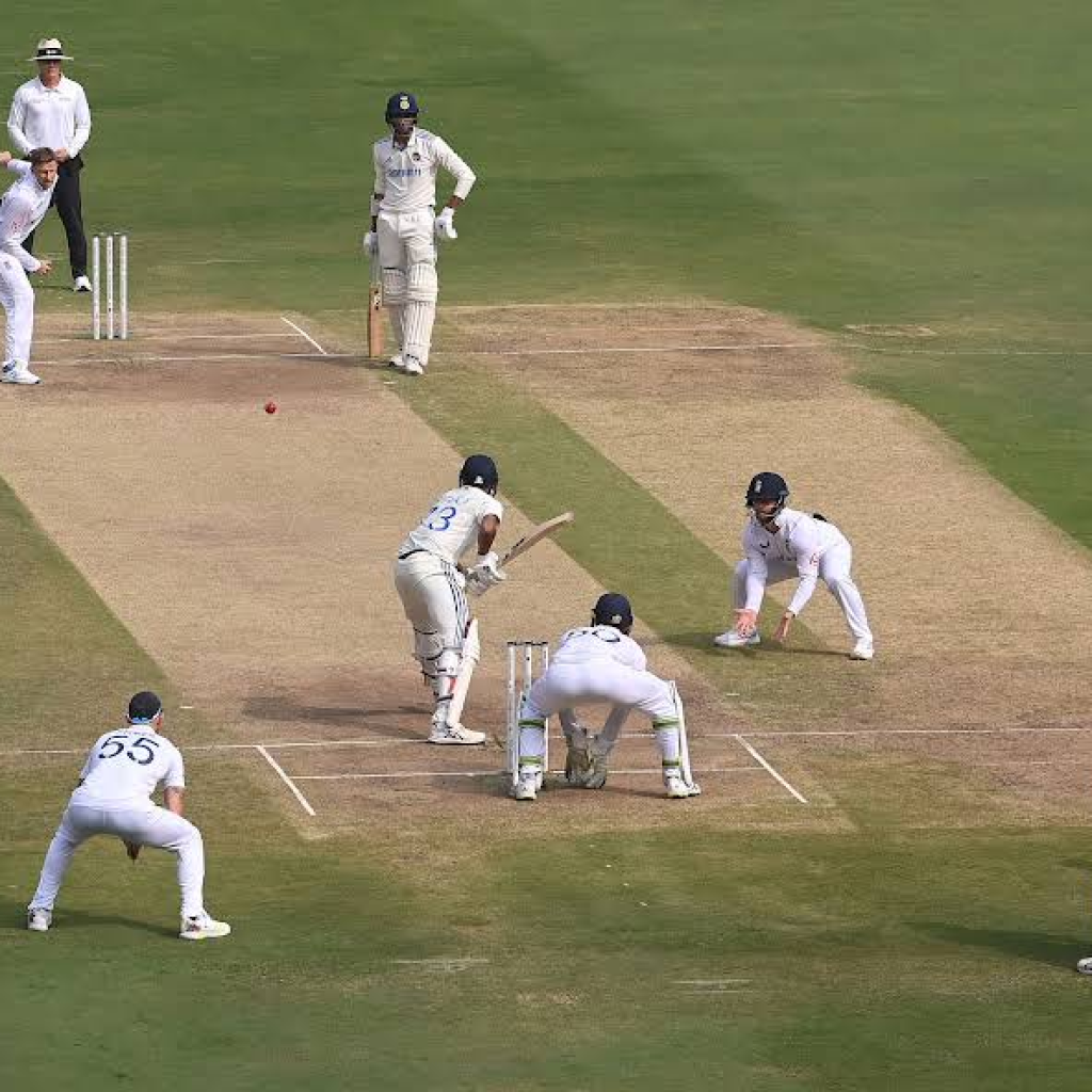 A bowler delivers to a batter on a cricket pitch with close fielders around the bat