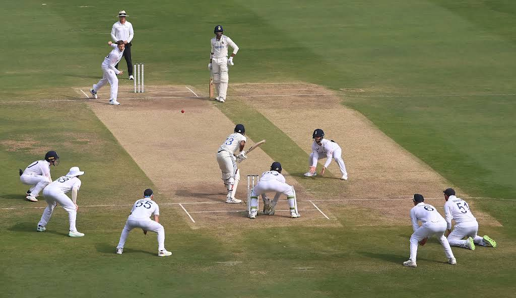 A bowler delivers to a batter on a cricket pitch with close fielders around the bat
